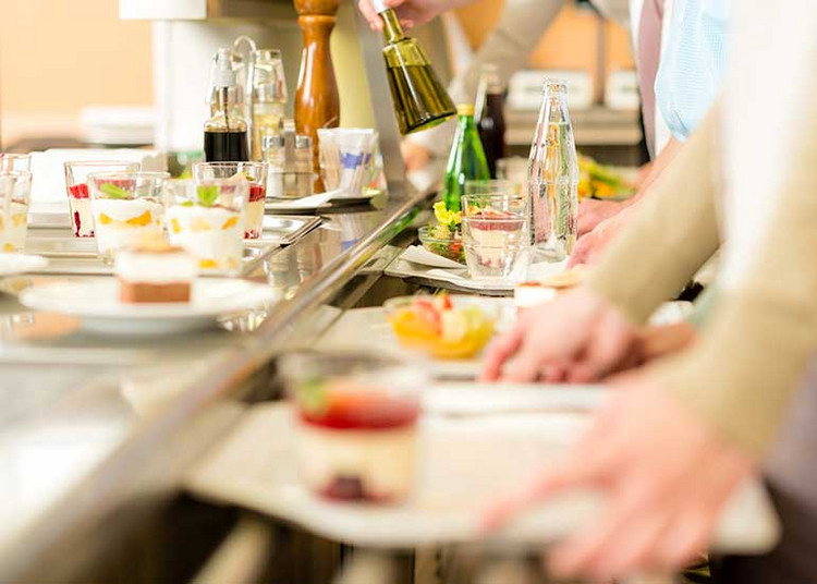 Personen stehen in einer Schlange zu einer Essensausgabe und stellen Gerichte auf ihr Tablet.