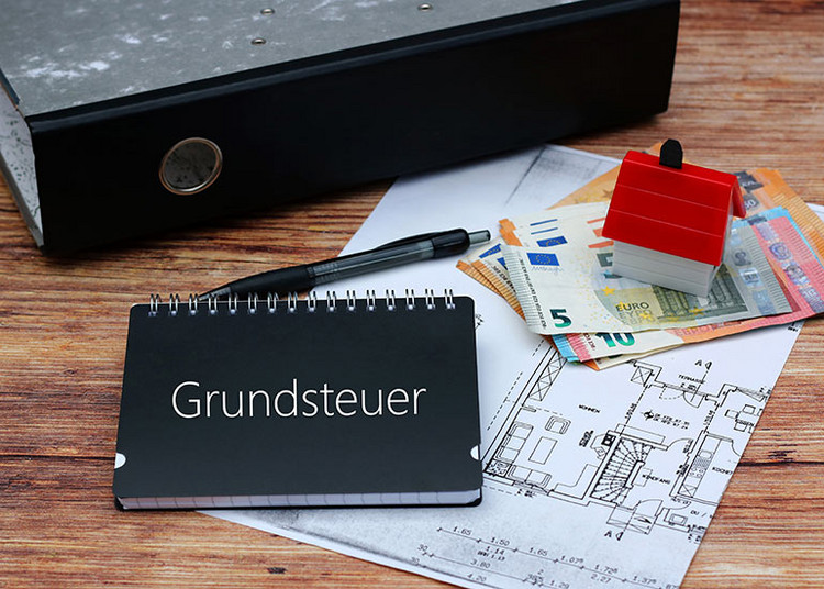 Auf dem Tisch liegt ein Bauplan. Darauf liegen Euro-Banknoten, ein Kugelschreiber, ein Aktenordner sowie ein Modell eines Hauses. Ebenso ein Notizbuch mit der Beschriftung „Grundsteuer”.