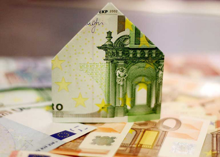 Auf einem Tisch liegen Euro-Banknoten. Auf den Geldscheinen ist eine Euro-Banknote zu sehen, die zu einem Haus gefaltet ist.