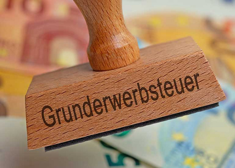 Auf einem Stempel steht „Grunderwerbsteuer”, dahinter sind Euro-Banknoten zu sehen.