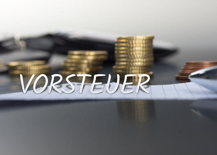 In einer Bildcollage ist der Schriftzug „Vorsteuer” vor einem Tisch platziert, auf dem ein Blatt Papier und ein Stapel Euromünzen liegen.