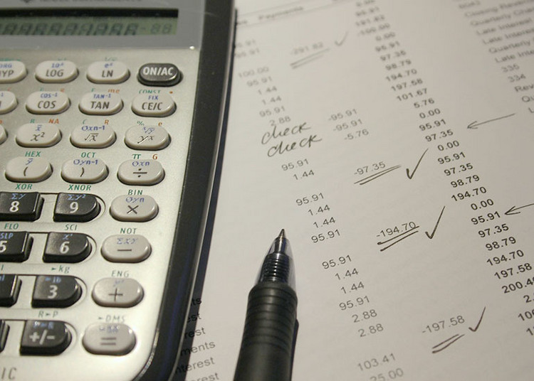 Neben einem Taschenrechner liegt ein Blatt Papier mit Zahlenspalten. Einzelne Zahlen wurden mit einem Stift als geprüft markiert. Der Stift liegt auf dem Papier. 