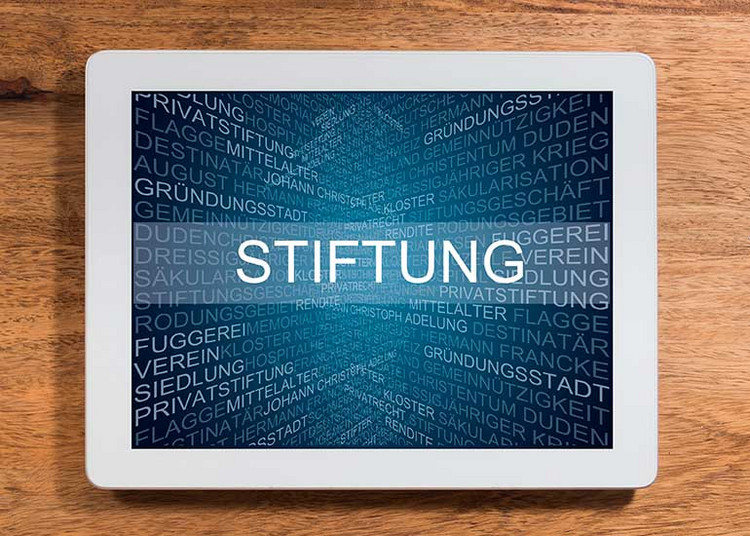 Auf einem Tablet ist der Text „Stiftung” geschrieben. Dahinter sind viele Begriffe wie Rendite, Privatrecht und Kloster angeordnet, die damit in Zusammenhang stehen.