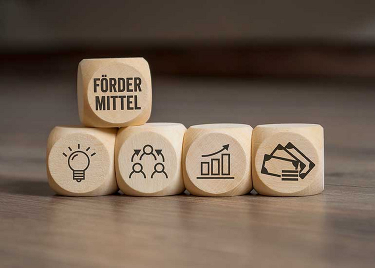 Auf einem Tisch liegen fünf Holzwürfel. In der untersten Reihe sind auf vier Würfeln Grafiken mit Abbildungen einer Glühbirne, Geldscheinen, einem aufsteigenden Balkendiagramm und drei Personen in einem Kreis zu sehen. Darüber liegt ein weiterer Würfel, auf dem „Födermittel” steht.