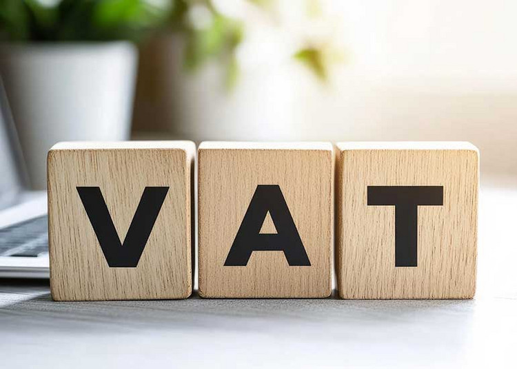 Die Buchstaben "VAT" für Mehrwertsteuer stehen auf drei Bauklötzen auf einem Schreibtisch.