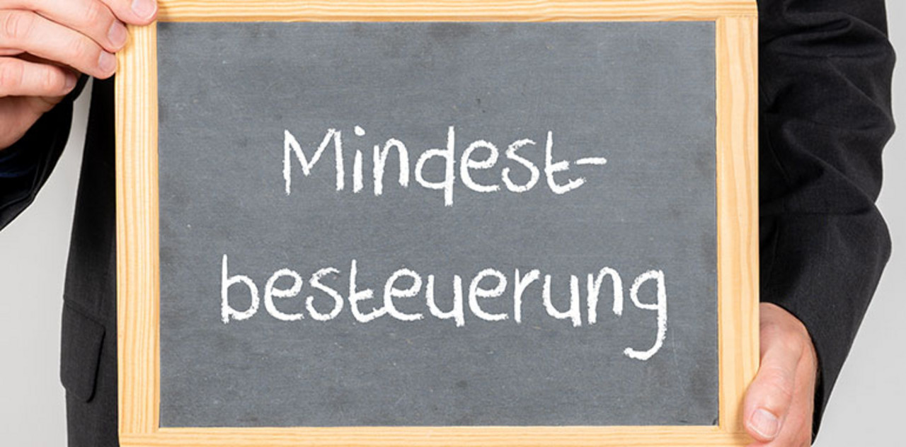 Person hält eine Tafel mit Aufschrift "Mindestbesteuerung" 