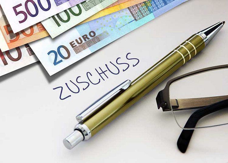 Auf einem Blatt Papier steht das Wort „Zuschuss”. Daneben liegen Euro-Banknoten, ein Kugelschreiber und eine Brille.