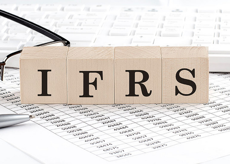 Bauklötze, die die Buchstaben "IFRS" bilden. Diese sind auf Blättern mit Zahlentabellen und einer Lesebrille angeordnet. Jeder Buchstabe steht auf einem eigenen Klotz, die zusammen den Begriff IFRS (International Financial Reporting Standards) darstellen.