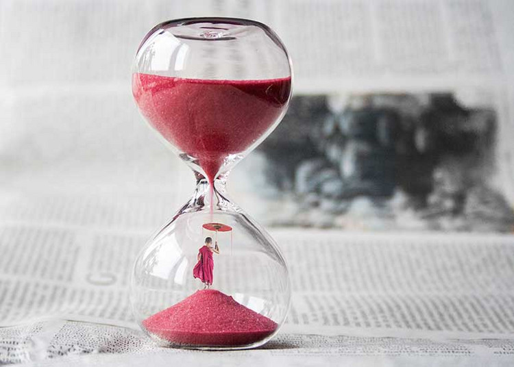 Auf einer aufgeschlagenen Zeitung steht eine Sanduhr, durch die roter Sand fließt.