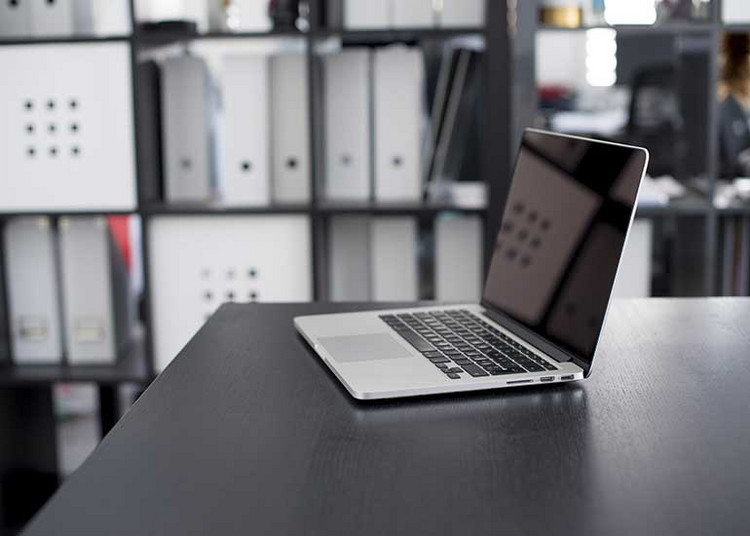 Ein Laptop steht auf einem Tisch. Im Hintergrund ist ein Schrank mit Aktenordnern zu sehen.