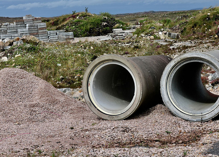Eine unbebaute, steinige Landschaft mit Baustoffen. Im Vordergrund liegen zwei große, graue Betonrohre auf einem Schotterhaufen.