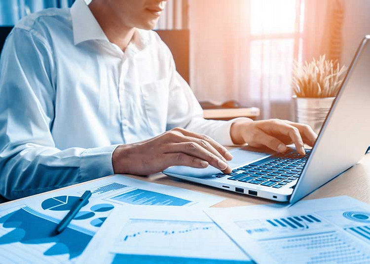 Person sitzt mit Laptop an Schreibtisch. Auf dem Schreibtisch liegen auch noch Blätter mit Grafiken.
