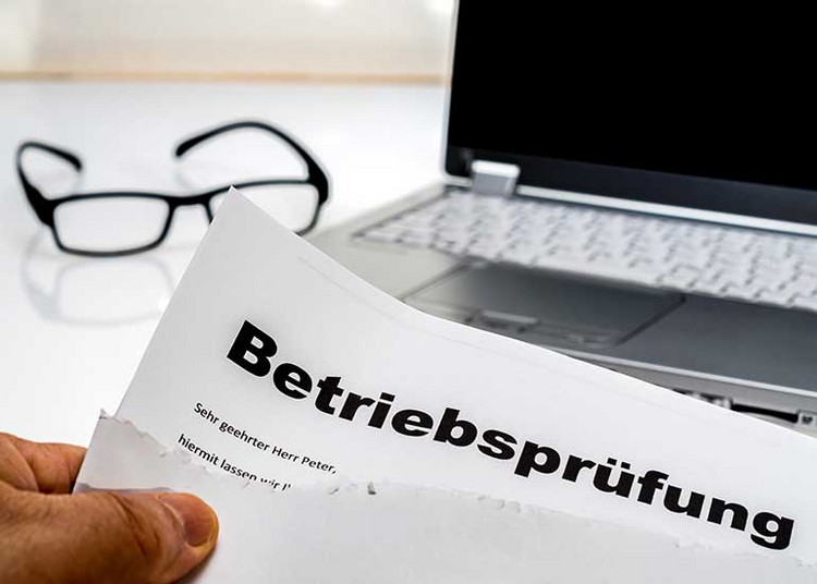 Eine Person hält ein Dokument mit der Überschrift „Betriebsprüfung” in der Hand. Im Hintergrund sind ein Laptop und eine Brille zu sehen.