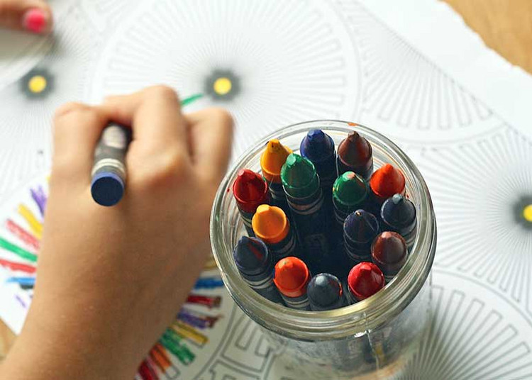 Ein Kind malt mit Wachsmalstiften in einem Malbuch. Auf dem Malbuch steht ein Glas mit vielen unterschiedlichen Stiften.
