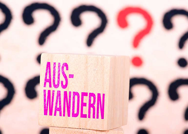 Auf einem Würfel steht "Auswandern". Im Hintergrund sind viele Fragezeichen zu sehen.