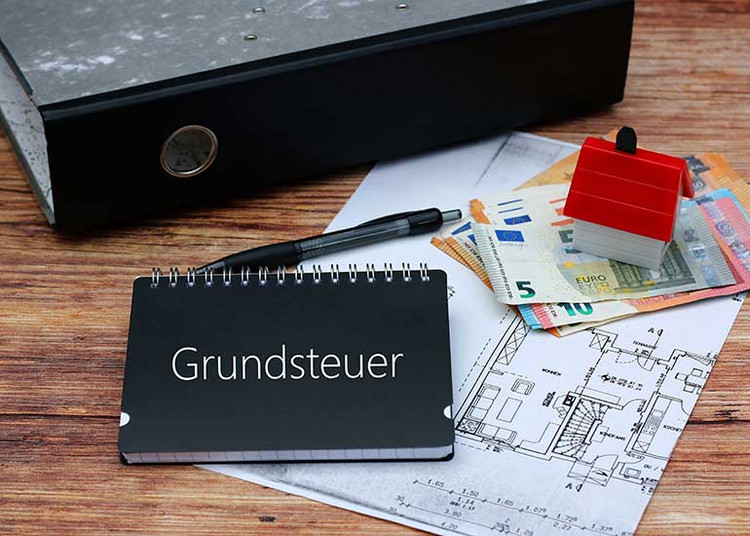 Auf dem Tisch liegt ein Bauplan. Darauf liegen Euro-Banknoten, ein Kugelschreiber, ein Aktenordner sowie ein Modell eines Hauses. Ebenso ein Notizbuch mit der Beschriftung „Grundsteuer”.