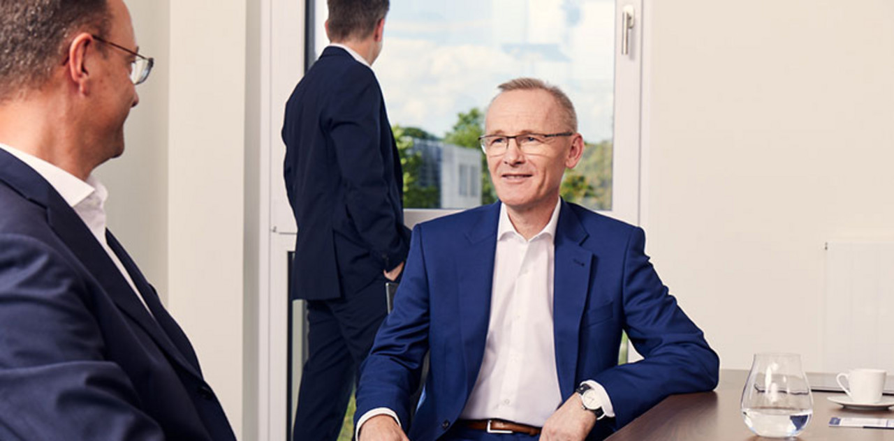Zwei Partner sitzen an einem Tisch und unterhalten sich. Ein weiterer steht im Hintergrund.