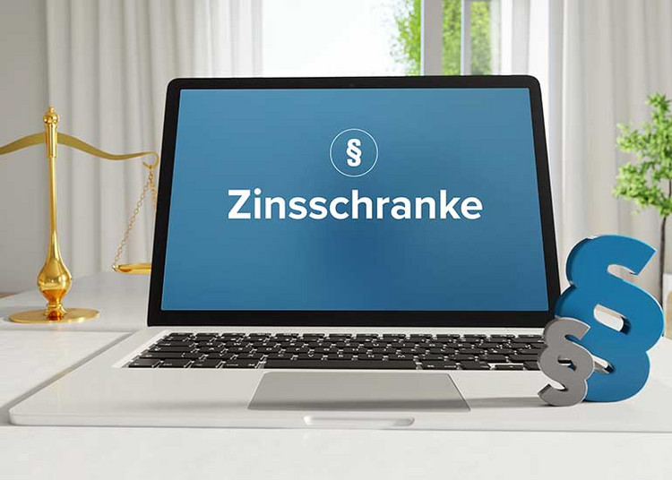 Auf einem Schreibtisch steht ein Laptop, auf dessen Bildschirm ein Paragraphenzeichen und das Wort "Zinsschranke" zu sehen sind. Im Hintergrund befindet sich eine Richterwaage und daneben zwei weitere Paragraphenzeichen.