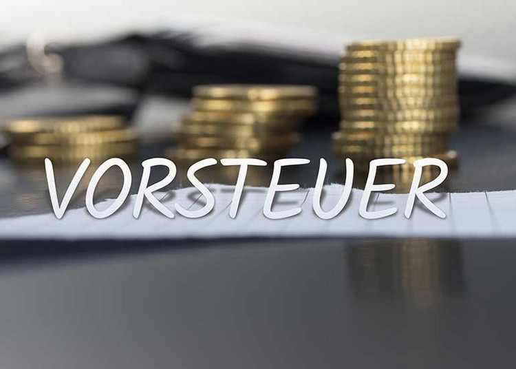 In einer Bildcollage ist der Schriftzug „Vorsteuer” vor einem Tisch platziert, auf dem ein Blatt Papier und ein Stapel Euromünzen liegen.