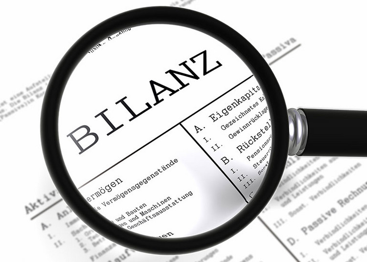 Eine Lupe vergrößert das Wort "Bilanz" auf einem Blatt Papier.