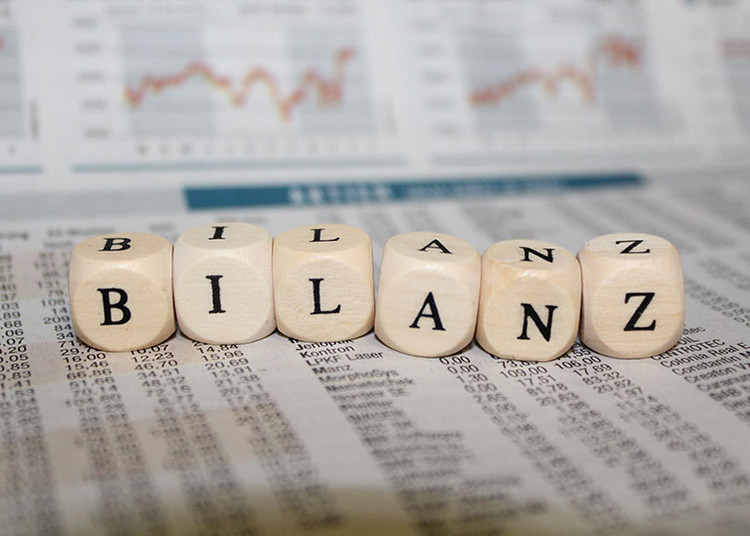 Holzbuchstaben bilden das Wort „Bilanz“ auf einem Finanzdokument mit Tabellen und Diagrammen im Hintergrund.