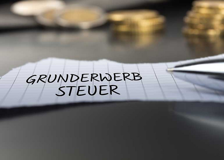Auf einem Tisch liegt ein Zettel, auf dem „Grunderwerbsteuer” steht. Im Hintergrund sind Stapel aus Münzen und daneben ein Kugelschreiber zu sehen.
