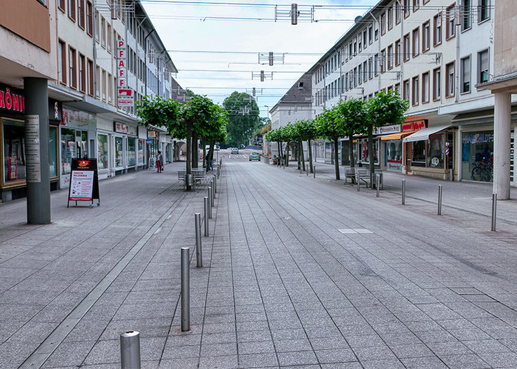 Breite, nahezu menschenleere Fußgängerzone in einer Stadt. Moderne Geschäftsgebäude und Schaufenster säumen beide Seiten der gepflasterten Straße. Gleichmäßig angeordnete Bäume, Sitzgelegenheiten und Poller strukturieren den öffentlichen Raum. Die Perspektive führt entlang der Einkaufsstraße in die Ferne unter einem leicht bewölkten Himmel.