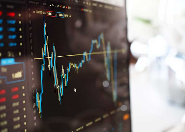 Nahaufnahme eines Computerbildschirms mit einem Finanz- und Börsendiagramm. Die Anzeige zeigt Kursverläufe und Schwankungen in Form von Linien und Kerzencharts auf dunklem Hintergrund.