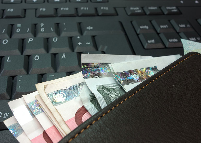 Auf einer Tastatur liegt ein Portemonnaie. Aus dem Portemonnaie ragen Euro-Banknoten.