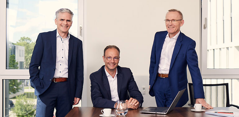 Drei Partner stehen an einem Schreitisch zusammen.