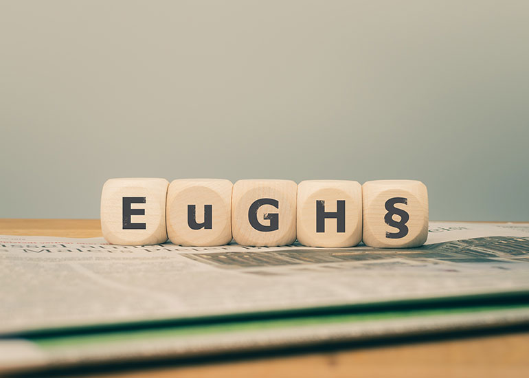 Auf Würfeln steht "EUGH" und ein "§". Das ist die Abkürzung für den Europäischen Gerichtshof. Die Buchstaben stehen einzeln auf den Würfeln. Unter den Würfeln liegt eine Zeitung auf einem Tisch.