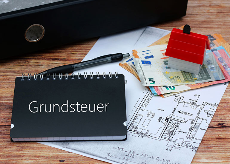 Auf einem Tisch liegt ein Aktenordner neben dem Grundriss eines Gebäudes. Auf dem Grundriss liegen außerdem Euro-Banknoten, das Modell eines Hauses, ein Kugelschreiber und ein Ringbuch mit der Aufschrift "Grundsteuer".