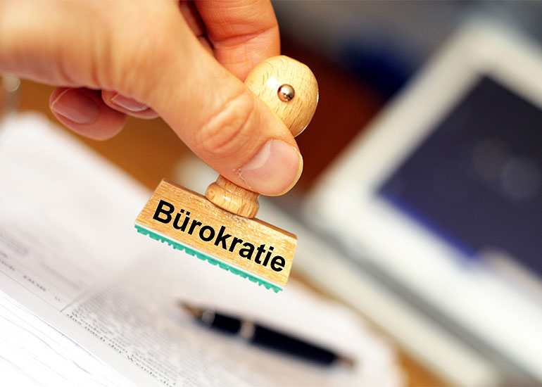Eine Hand hält einen Stempel mit der Aufschrift "Bürokratie". Im Hintergrund ist unscharf ein Schreibtisch mit Papieren zu erkennen.