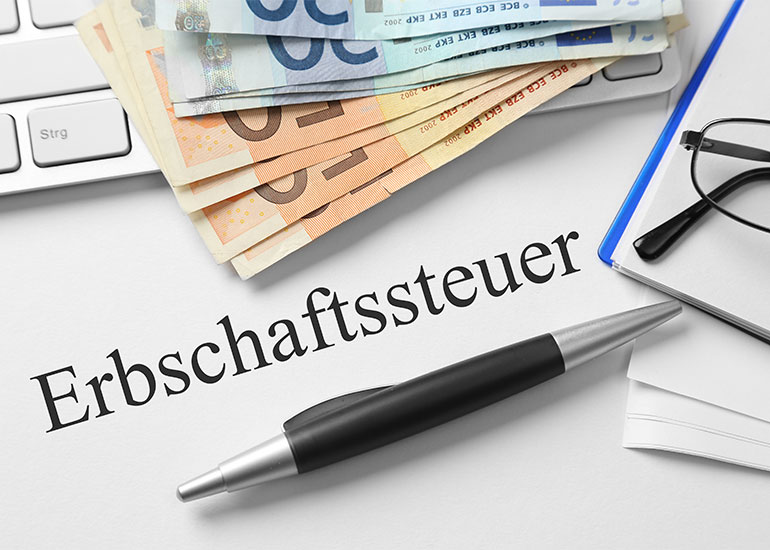 Auf einem Zettel steht "Erbschaftssteuer". Daneben liegen Euroscheine, ein Kugelschreiber, eine Tastatur und eine Brille.