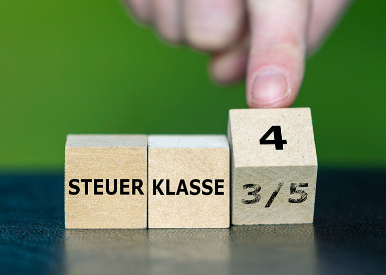 Auf einem Tisch liegen drei Würfel. Auf dem ersten steht "Steuer", auf dem zweiten "Klasse" und auf dem dritten wird mit dem Finger "3/5" in "4" geändert.