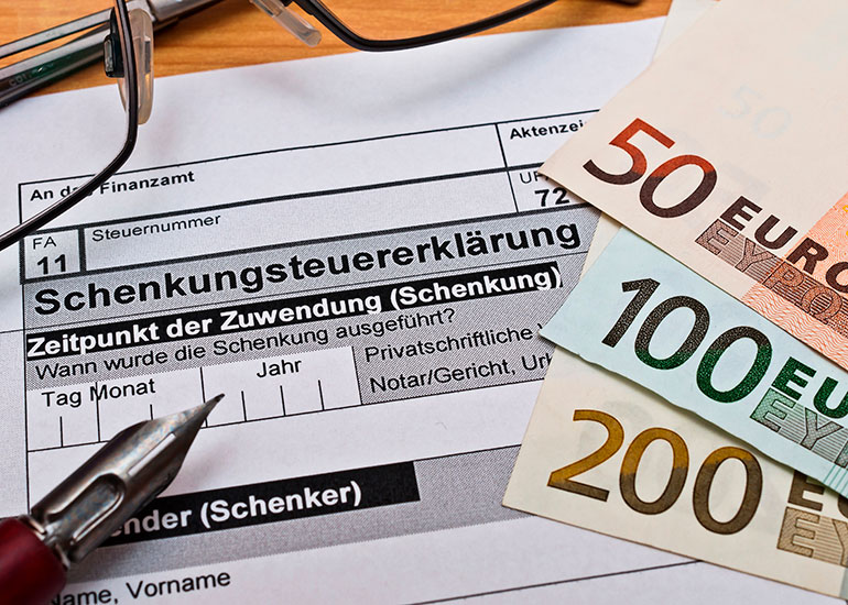 Auf einem Tisch liegt ein Formular für eine Schenkungsteuererklärung. Daneben liegen eine Brille, ein Kugelschreiber und Euroscheine.