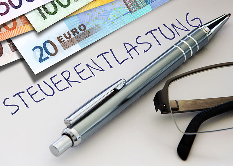 Auf einem Papier steht „Steuerentlastung”. Darauf liegen Euro-Banknoten, ein Kugelschreiber und eine Brille.