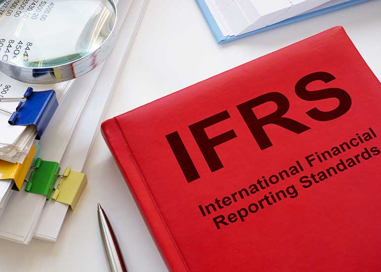 Auf einem Tisch liegt ein Buch mit der Aufschrift „IFRS – International Reporting Standards”. Daneben liegen ein weiteres Buch, ein Kugelschreiber und einige geheftete Blätter Papier, auf denen eine Lupe liegt.