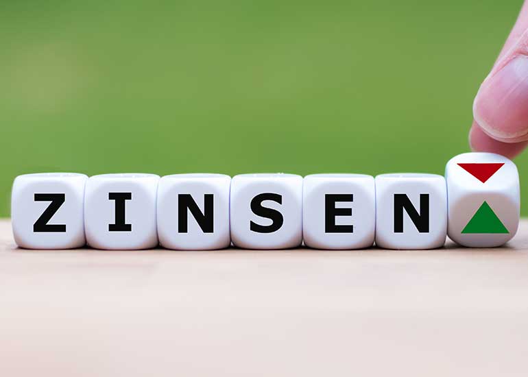 Das Wort „Zinsen” ist aus Würfeln geschrieben. Jeder Buchstabe steht auf einem eigenen Würfel. Eine Hand dreht einen weiteren Würfel um, sodass statt eines Pfeils nach oben ein Pfeil nach unten erscheint.