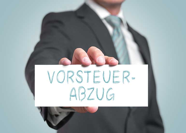 Eine Person im Anzug hält ein Schild mit der Aufschrift „Vorsteuerabzug”.