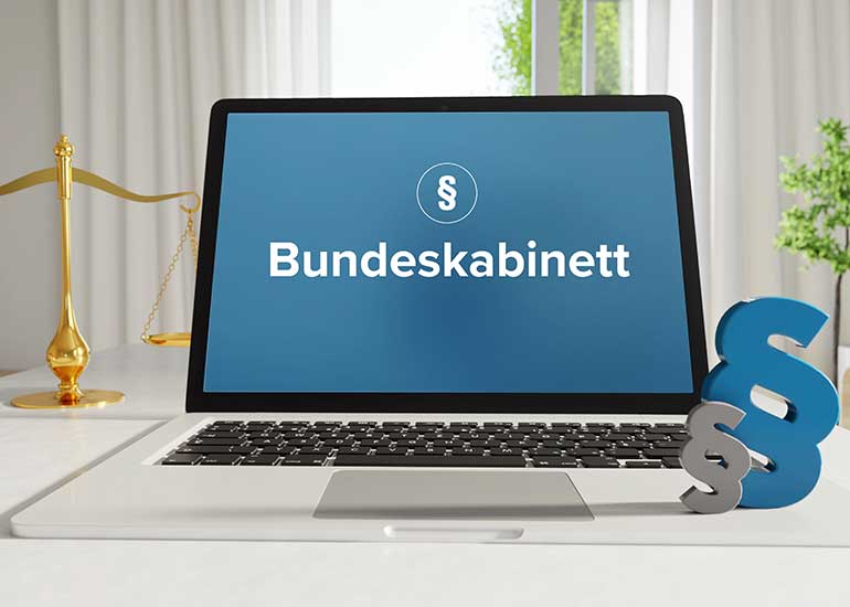 Auf einem Schreibtisch steht ein Laptop, auf dessen Bildschirm ein Paragraphenzeichen und das Wort "Bundeskabinett" zu sehen sind. Im Hintergrund befindet sich eine Richterwaage und daneben zwei weitere Paragraphenzeichen.