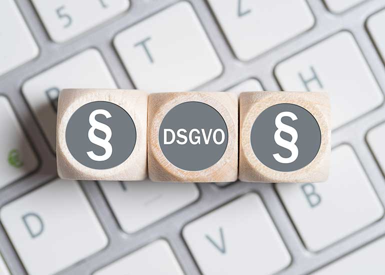 Auf einer Tastatur liegen drei Würfel in einer Reihe. Auf den beiden äußeren ist ein „§“-Zeichen abgebildet, auf dem Würfel in der Mitte steht „DSGVO“ für „Datenschutz-Grundverordnung“.