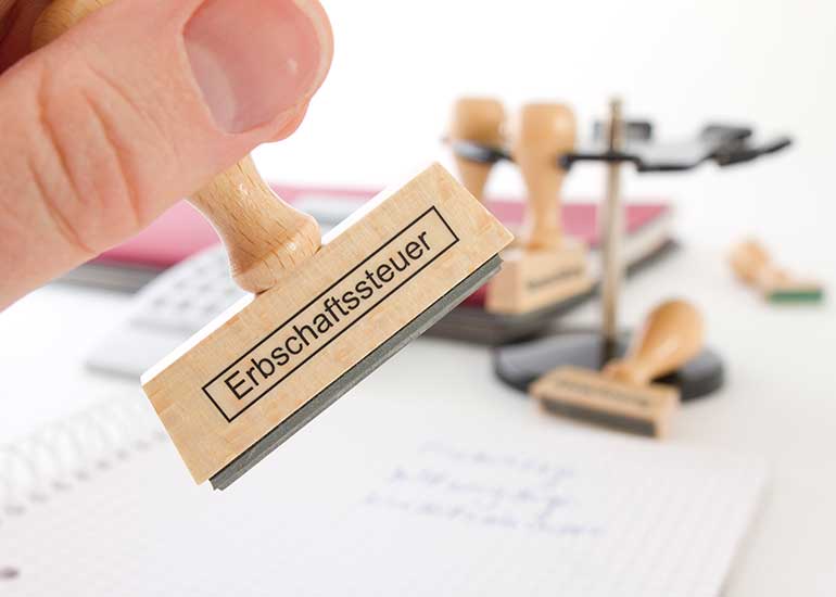 Die Person hält einen Stempel mit der Aufschrift „Erbschaftssteuer” in der Hand. Im Hintergrund ist ein Schreibtisch mit weiteren Würfeln zu sehen.