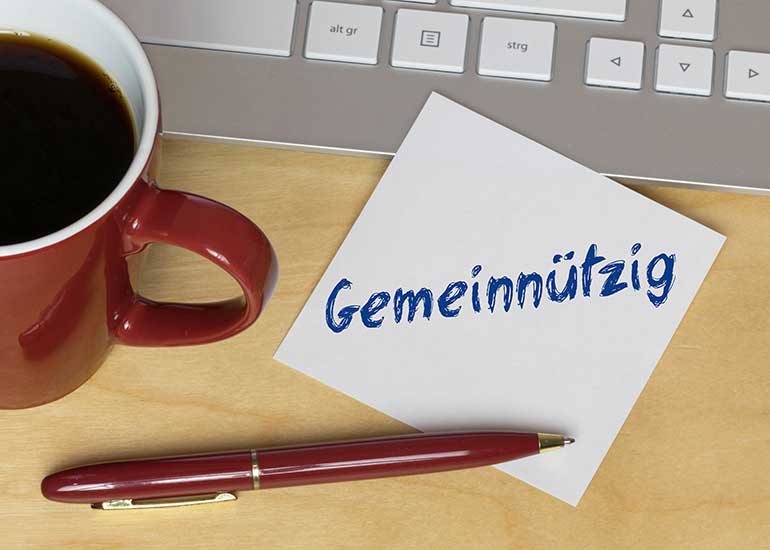 Auf dem Tisch stehen eine Tastatur, eine Kaffeetasse, ein Kugelschreiber und ein Zettel. Auf dem Zettel steht das Wort „Gemeinnützig”.