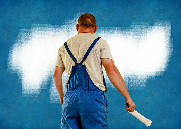 Zu sehen ist eine Person in Arbeitskleidung, die mit einer Malerrolle vor einer teilweise angestrichenen Wand steht.