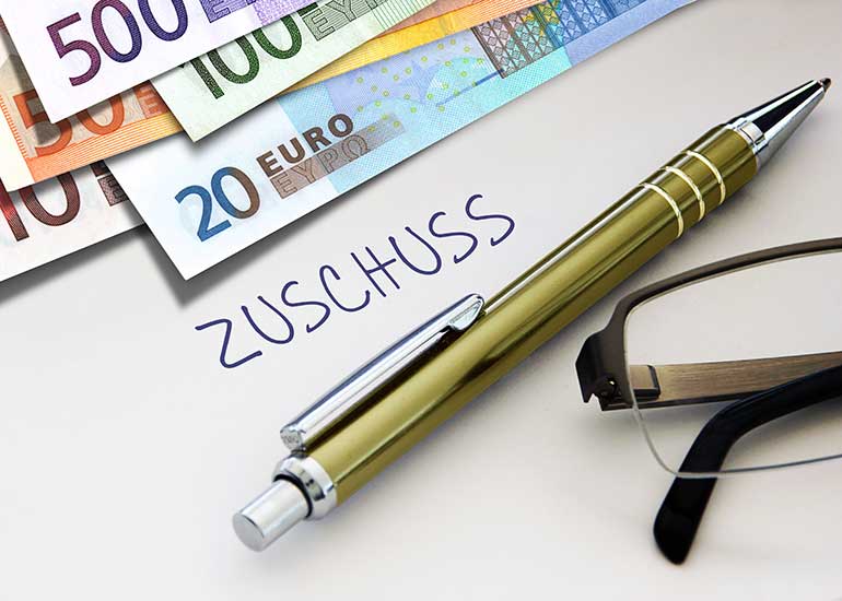 Auf einem Blatt Papier steht das Wort „Zuschuss”. Daneben liegen Euro-Banknoten, ein Kugelschreiber und eine Brille.