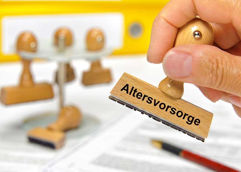 Eine Person hält einen Stempel in der Hand, auf dem „Altersvorsorge” steht. Im Hintergrund ist ein Schreibtisch zu sehen, auf dem sich weitere Stempel, bedruckte Blätter, ein Aktenordner und ein Stift befinden.