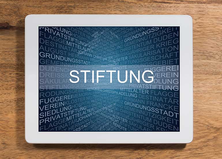 Auf einem Tablet ist der Text „Stiftung” geschrieben. Dahinter sind viele Begriffe wie Rendite, Privatrecht und Kloster angeordnet, die damit in Zusammenhang stehen.
