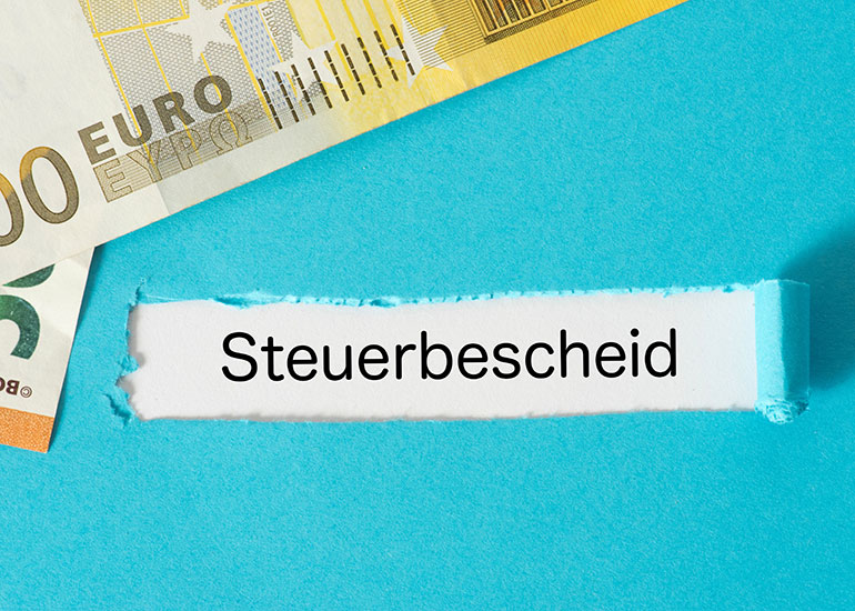 Hinter einem Loch in einem Papier wird der Schriftzug "Steuerbescheid" sichtbar. Daneben liegen Euro-Banknoten.