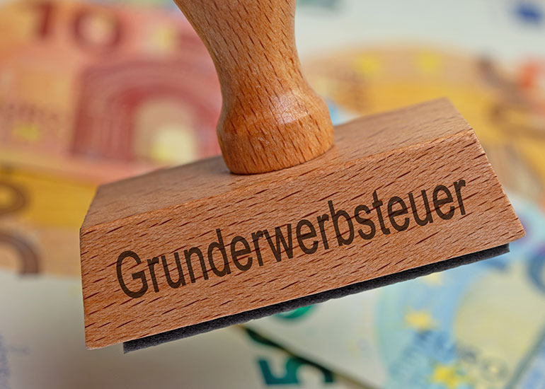 Auf einem Stempel steht „Grunderwerbsteuer”. Dahinter sind unscharf Euro-Banknoten zu erkennen.